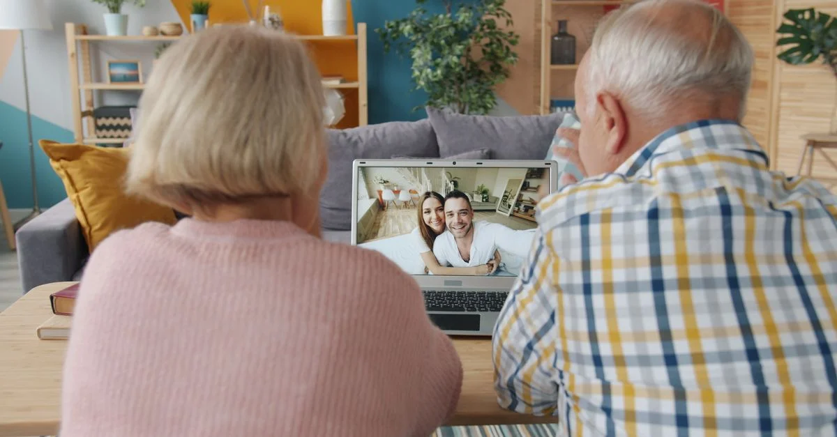 Un couple de personnes âgées profite d'un appel vidéo avec des membres de leur famille sur un ordinateur portable, confortablement installés dans leur salon.