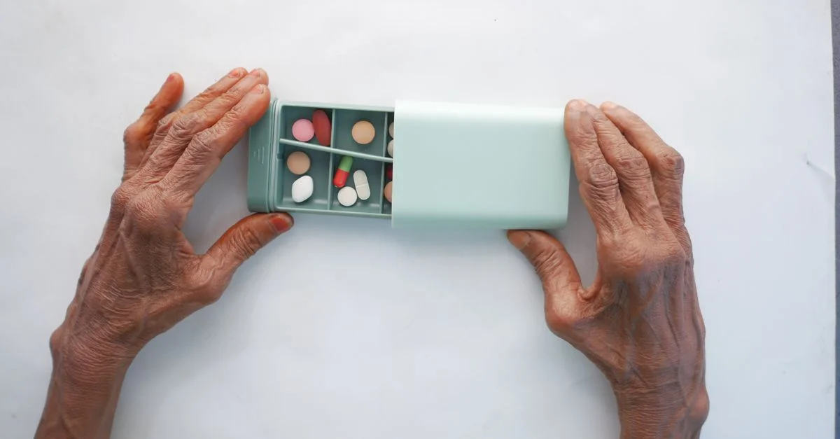 Personne âgée triant des pilules dans une boîte à pilules turquoise sur une surface blanche.