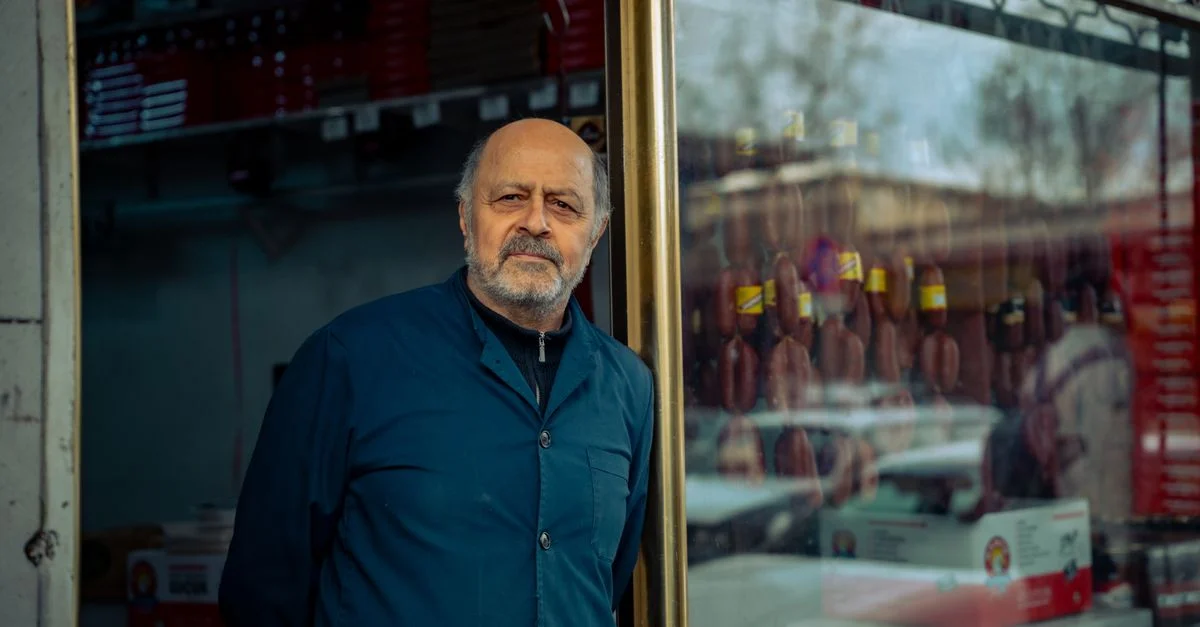 Un homme âgé se tient devant la vitrine de son magasin dans une zone urbaine, faisant la promotion d'un commerce local.