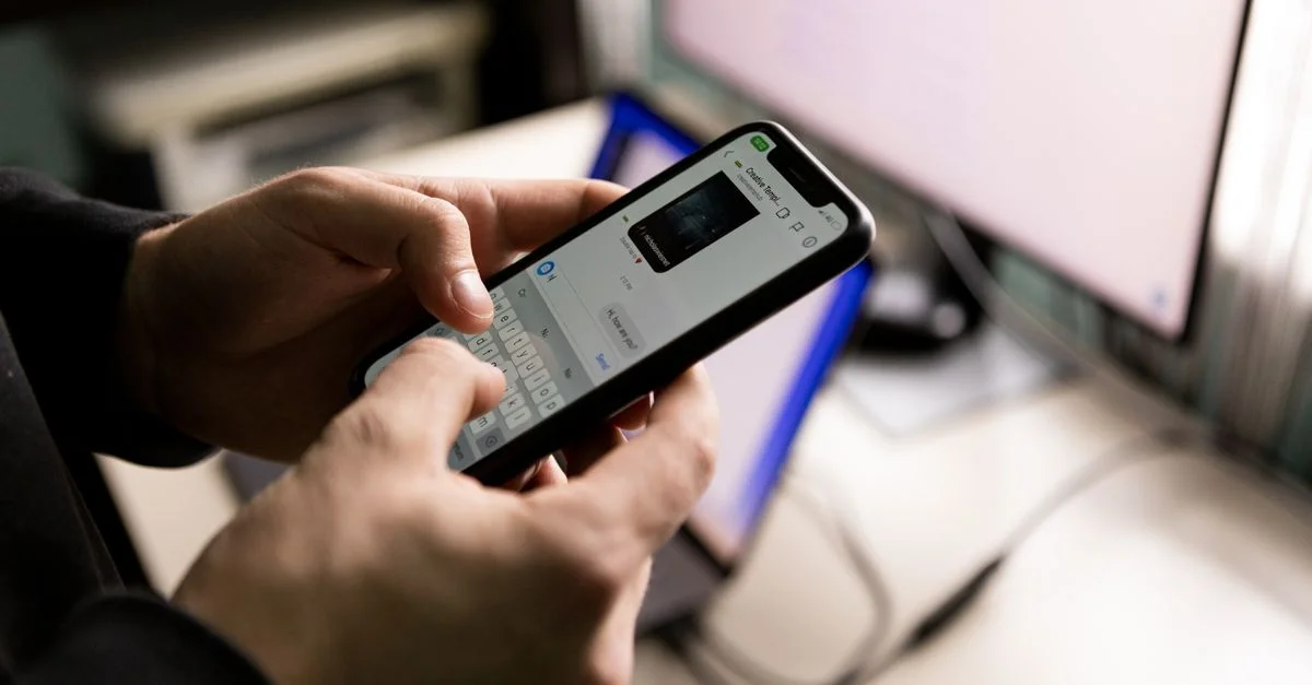 Une personne tenant un smartphone et discutant à l'intérieur près d'un écran d'ordinateur.